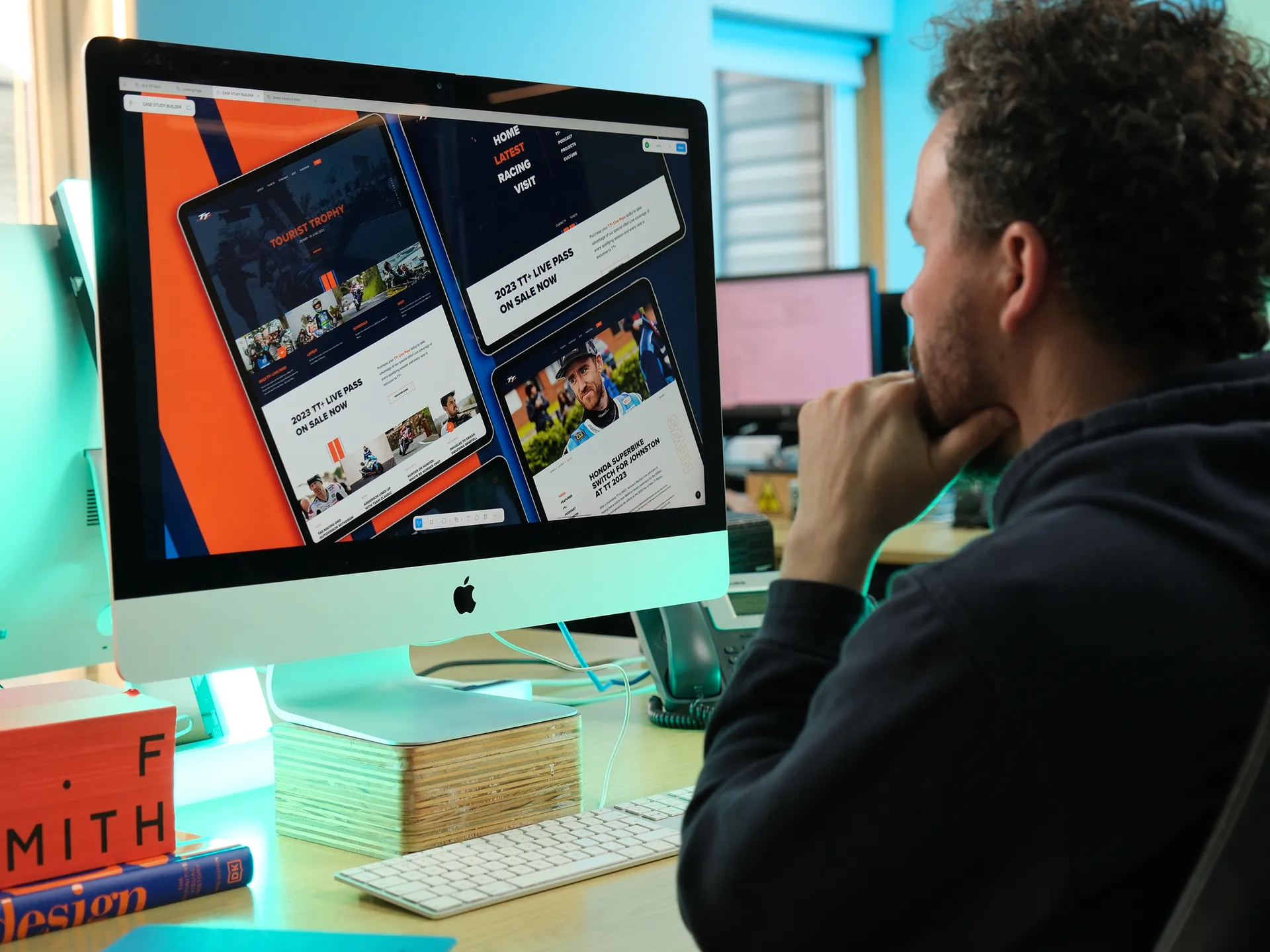Adrian Crellin reviewing the Isle of Man TT Races website iomttraces.com on an iMac at the Douglas office, with multiple page layouts visible on screen including the Tourist Trophy homepage and TT+ Live Pass page